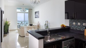 A modern kitchen with a black countertop, stainless steel sink, and a built-in dishwasher. There is a dining area with a white sofa and a chandelier.
