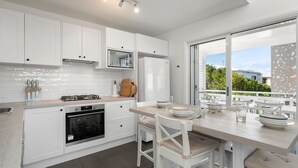 A modern kitchen with white cabinetry, a gas stove, an oven, and a dining area with a table and chairs. - By the Bay on Duncan by Experience Jervis Bay (Vincentia)