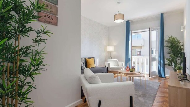 A modern living room with a sofa, armchair, and a dining table. There are plants, a lamp, and a view of the outside through large windows. - Stylish Comfortable Flat w/ Balcony by HostWise (Porto)