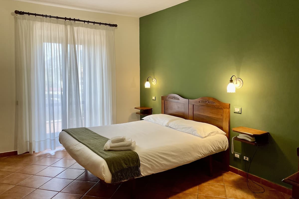 A hotel room with a bed, green walls, a wooden headboard, two bedside tables with lamps, a telephone, and a window with sheer curtains.
