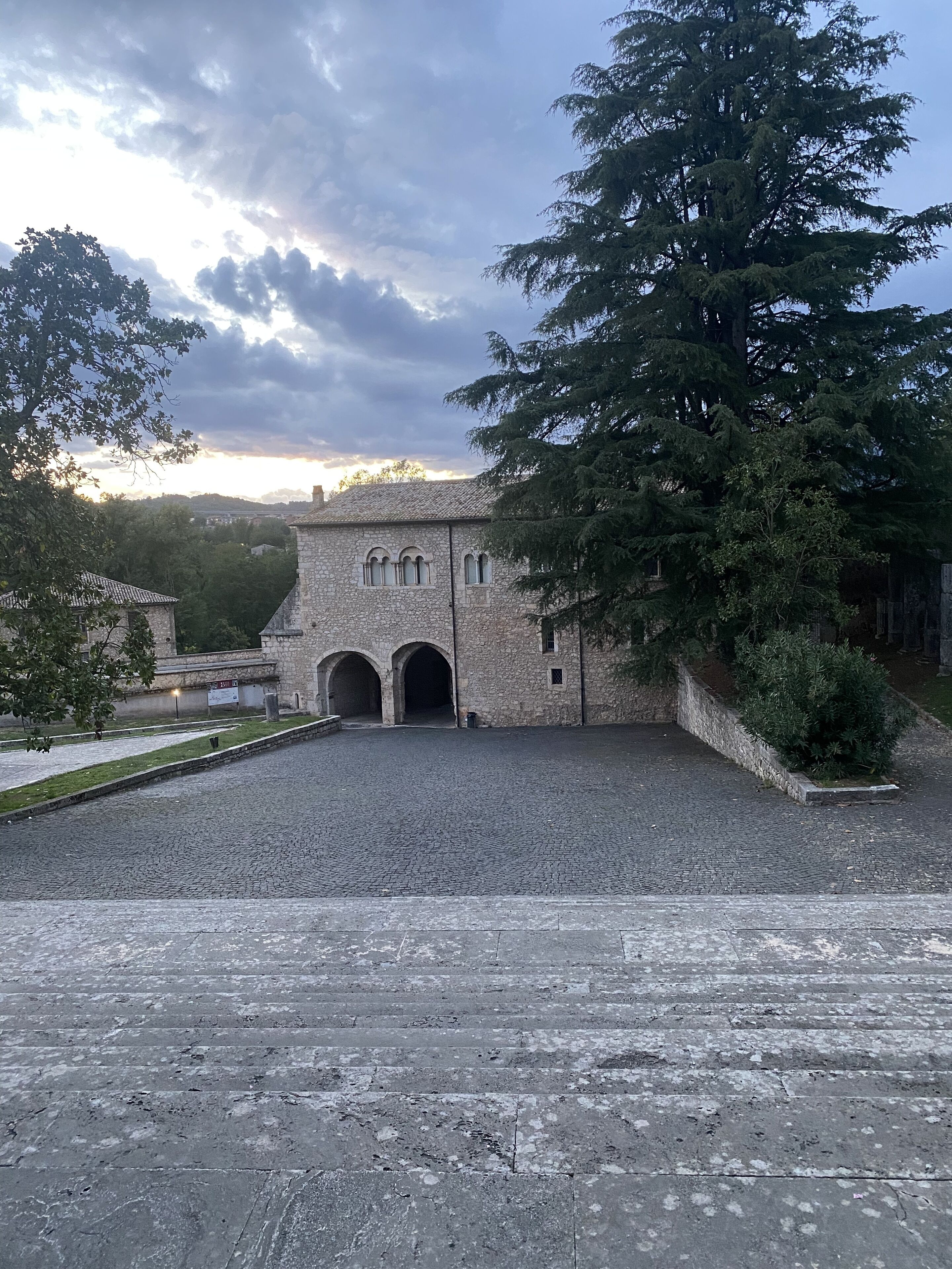 A stone building with arched entrances, a paved courtyard, and surrounding trees.