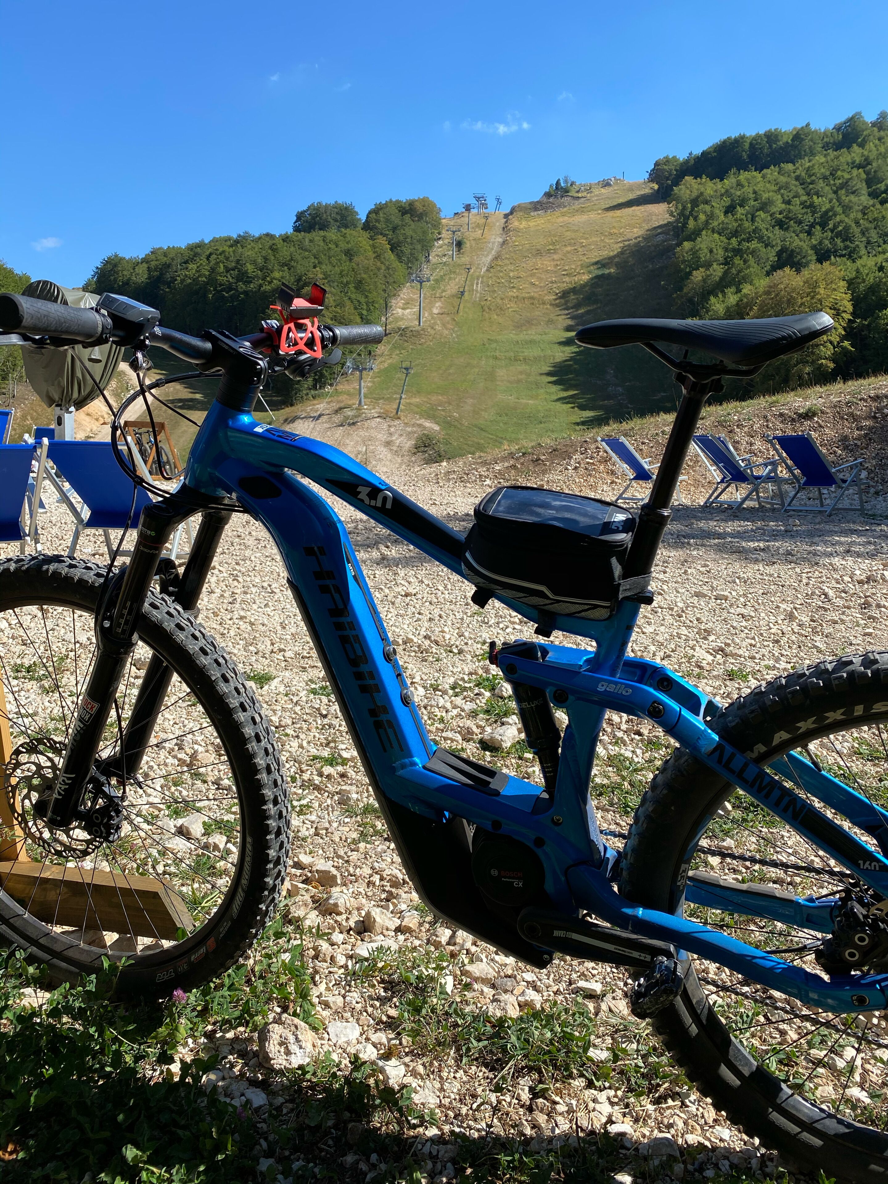 Un vélo électrique bleu est garé sur un chemin de gravier, offrant une vue pittoresque sur une colline et des arbres en arrière-plan.