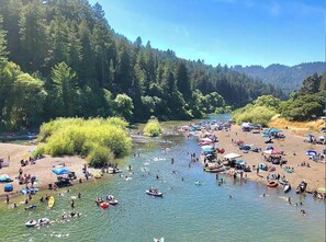 Un río con gente practicando actividades acuáticas y una zona de playa con sombrillas y tiendas de campaña.