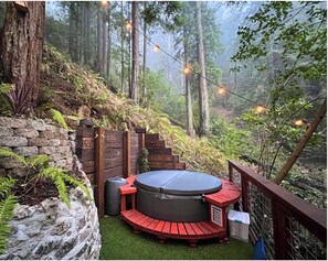 Un bain à remous sur une terrasse en bois, entouré d’un mur de pierres, avec une forêt en arrière-plan et des guirlandes lumineuses.