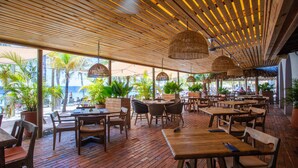 An outdoor restaurant with wooden tables and chairs, potted plants, and a wooden ceiling with hanging lights. - Blue Bay - The Reef (Sint Michiel)