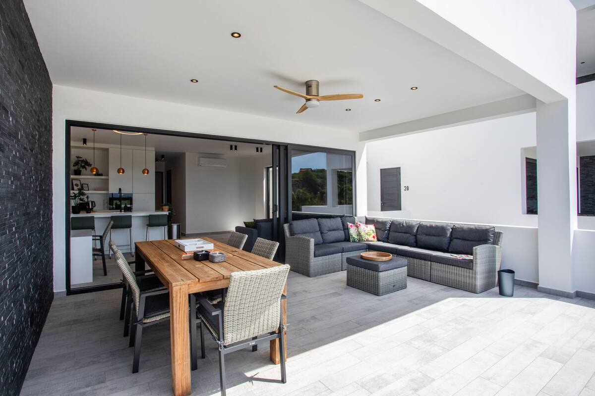 Un espacio exterior moderno con una mesa de comedor de madera, sillas de mimbre, un sofá gris y un ventilador de techo.