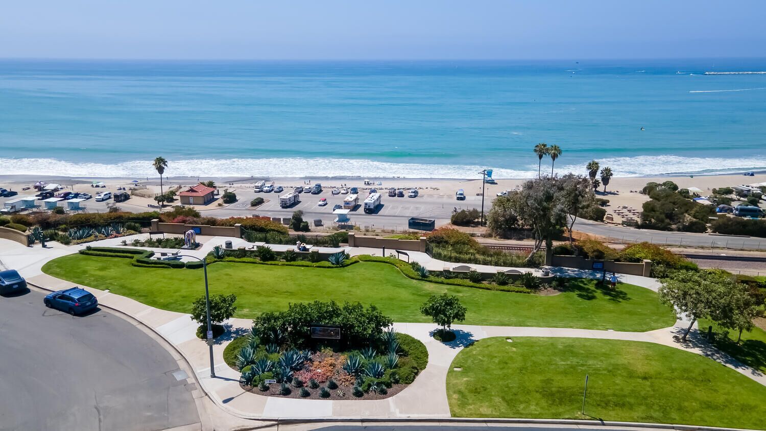 An aerial view of a coastal area with a parking lot, green lawns, and a beach in the background.