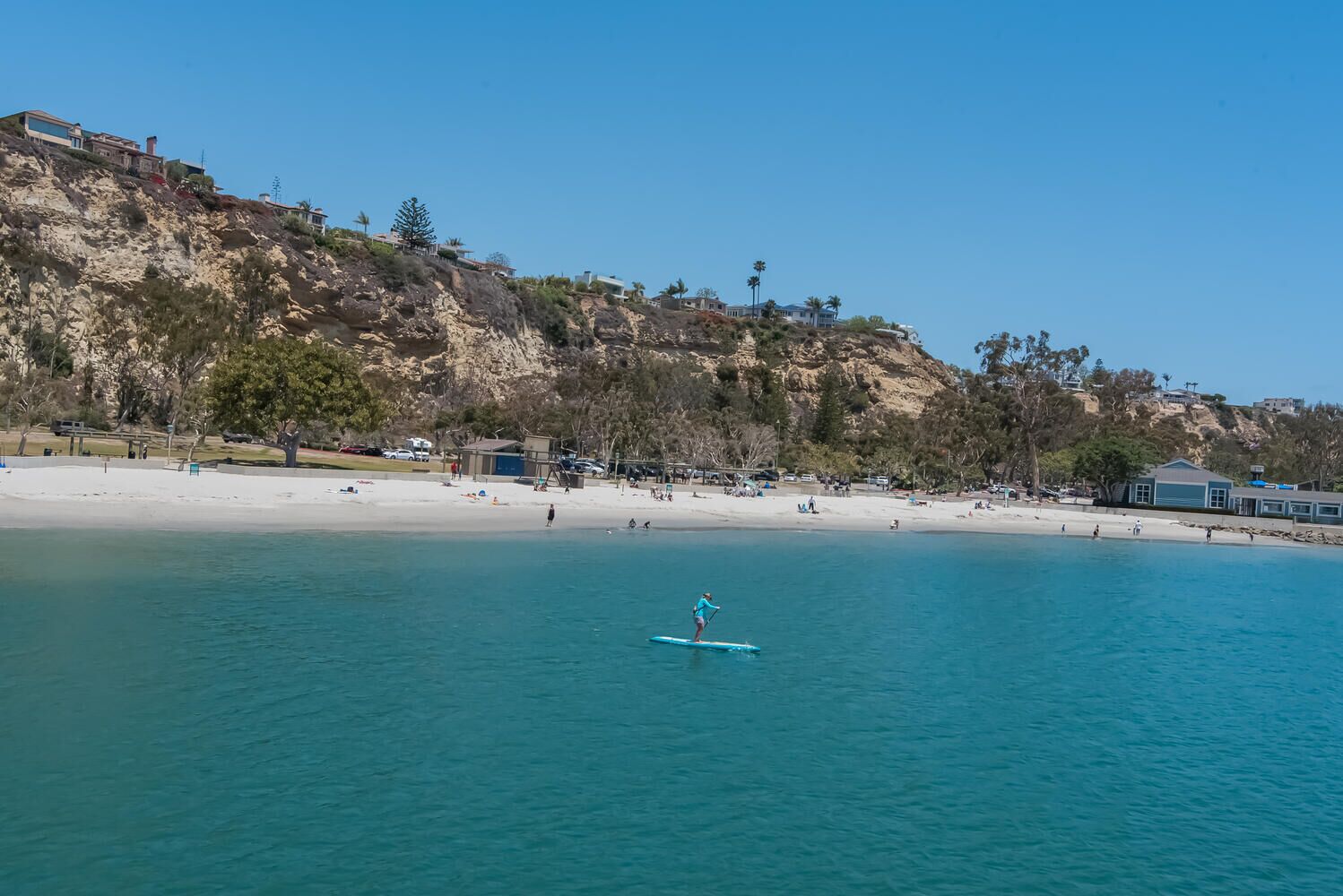 Une plage où l’on voit une personne faire du paddle, une eau d’un bleu limpide et, au loin, des bâtiments sur une colline.