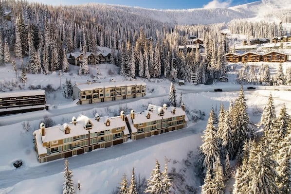 Uma cabana coberta de neve, com vários edifícios, rodeada por uma floresta densa e montanhas.