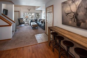 Uma sala de estar espaçosa com piso de madeira, um conjunto de sofás, uma mesa de centro e um espaço bar com banquetas.