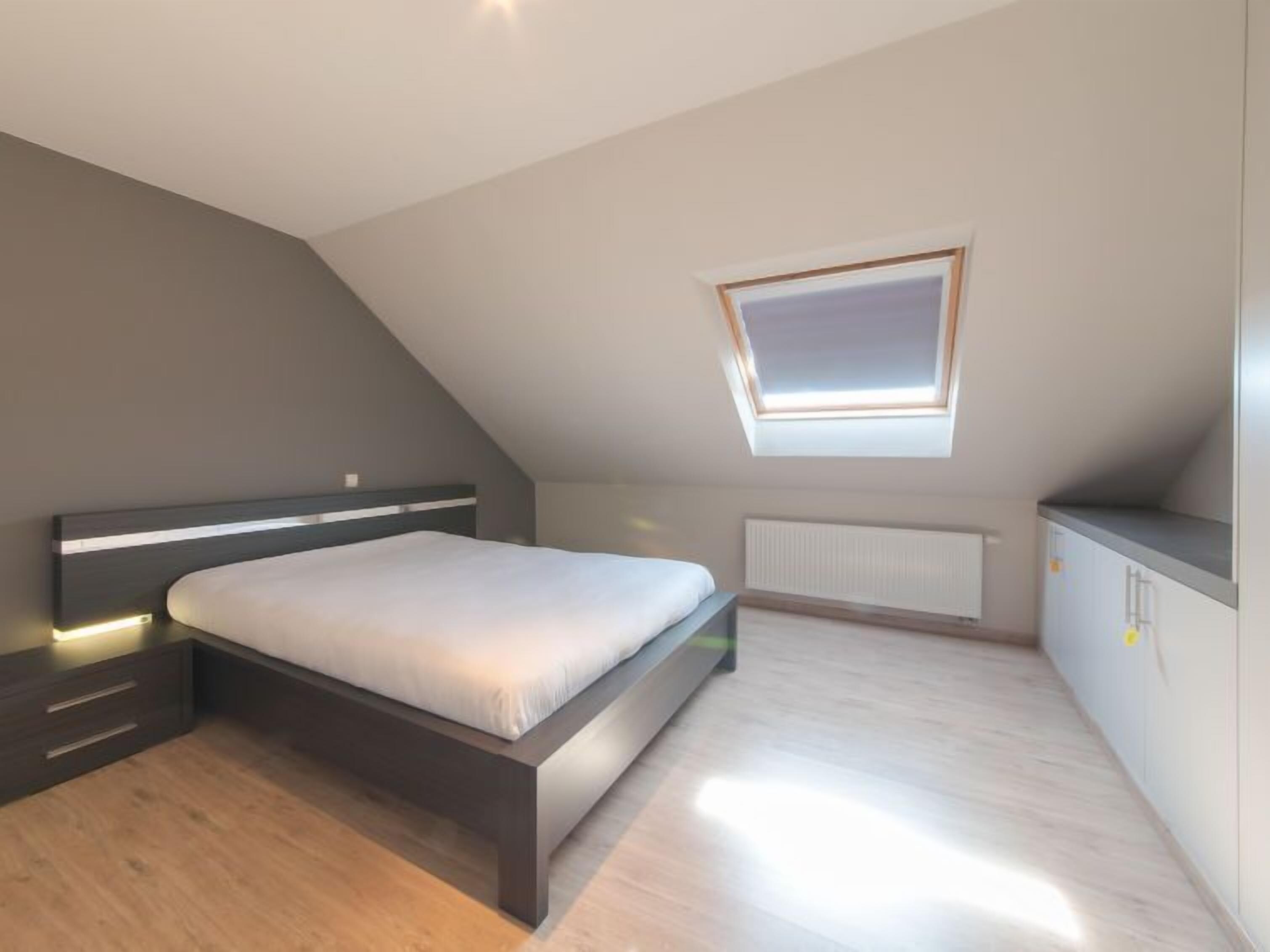 Un dormitorio moderno con techo inclinado, tragaluz, cama, mesita de noche y armario empotrado.