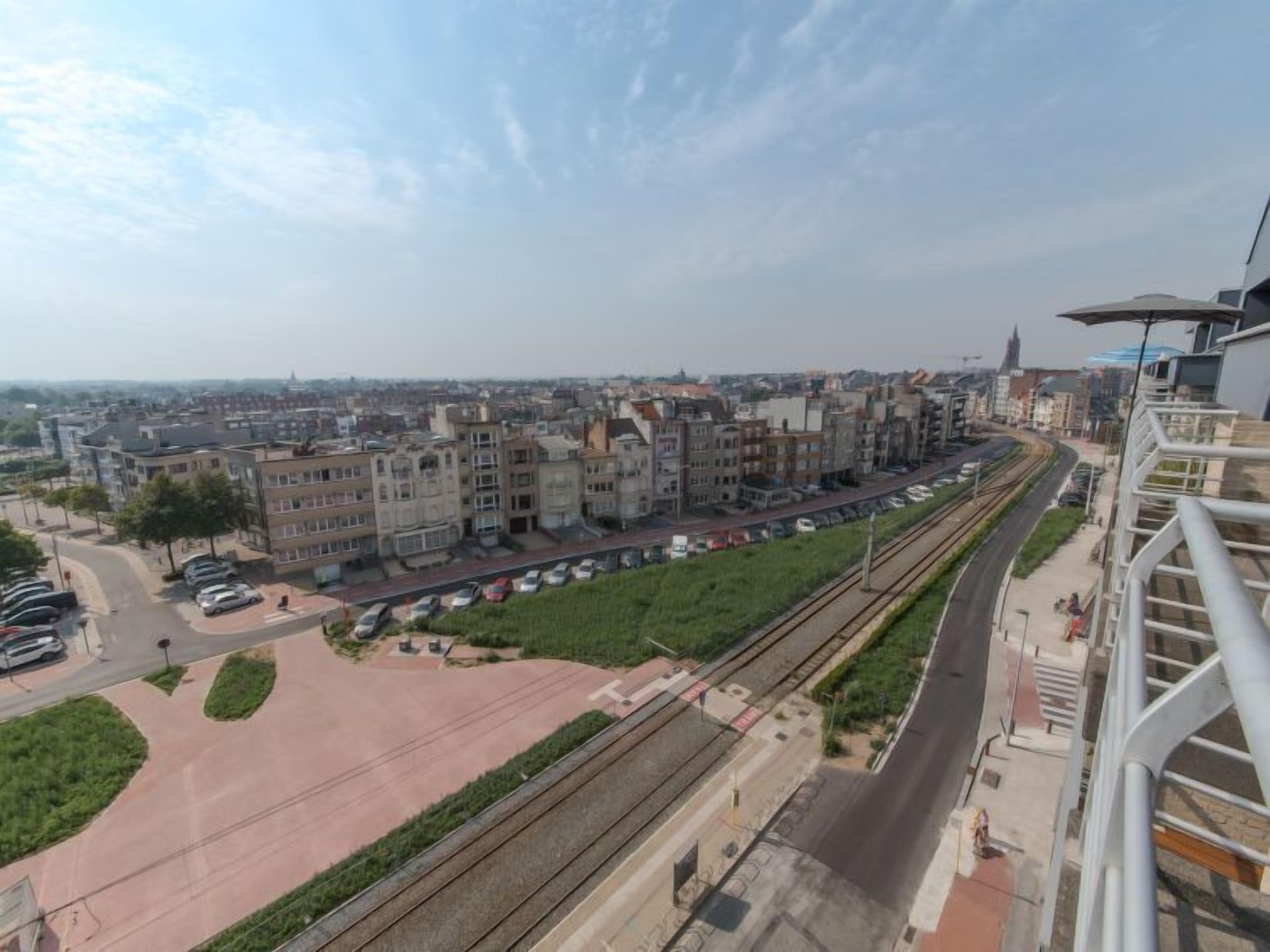 A high-rise building with a view of a cityscape, including a train track and parked cars.