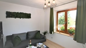 Een moderne woonkamer met een grijze bank, een klein tafeltje met een fles en glazen, een groene plant en een raam met gordijnen.