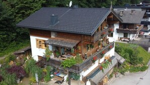 Een houten chalet met twee verdiepingen en een balkon, omgeven door weelderige begroeiing en een steile oprit.