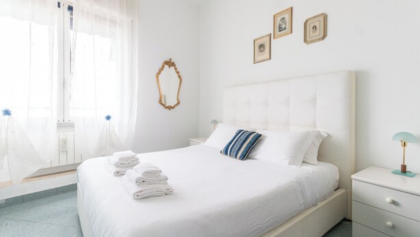 A hotel room with a large bed, a nightstand, a wall-mounted mirror, and a window with sheer curtains. - Italianway - Roma 70 (Sanremo)