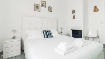 Een slaapkamer met een wit bed, nachtkastje, lamp, stoel en een klein kastje.