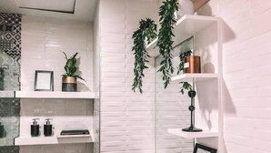 A bathroom with white tiled walls, a toilet, a sink with two soap dispensers, and shelves with plants and a picture frame.