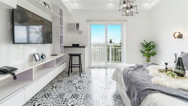 A modern bedroom with a large TV, a desk, a chair, a patterned floor, a balcony with plants, and a hanging light fixture.