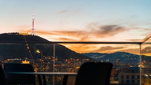 Un paisaje urbano al anochecer con una torre destacada en una colina, una vista a través de una ventana con sillas, y una cadena montañosa distante.