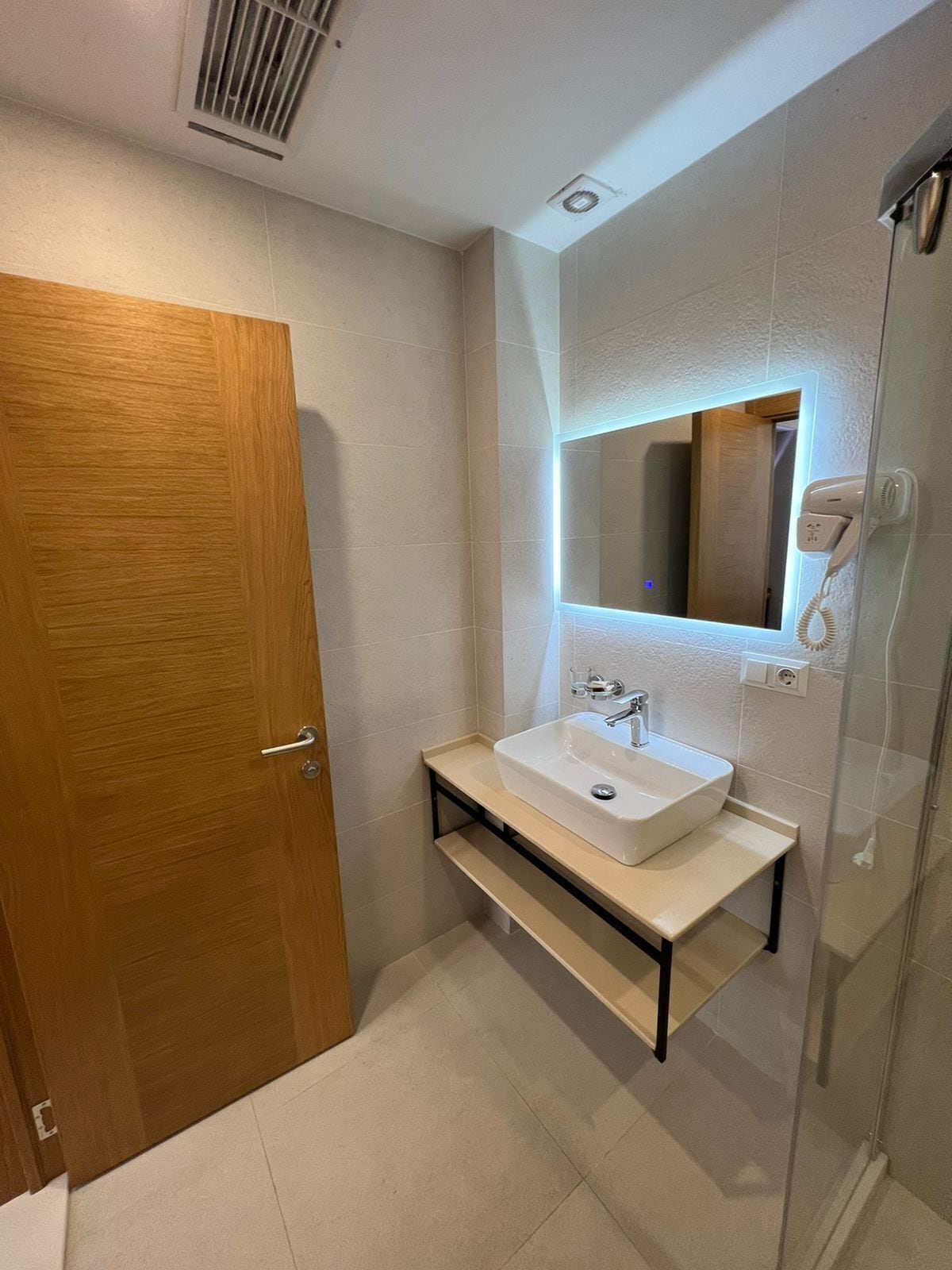 Une salle de bain moderne avec une porte en bois, un lavabo blanc surmonté d’un miroir et une cabine de douche en verre.