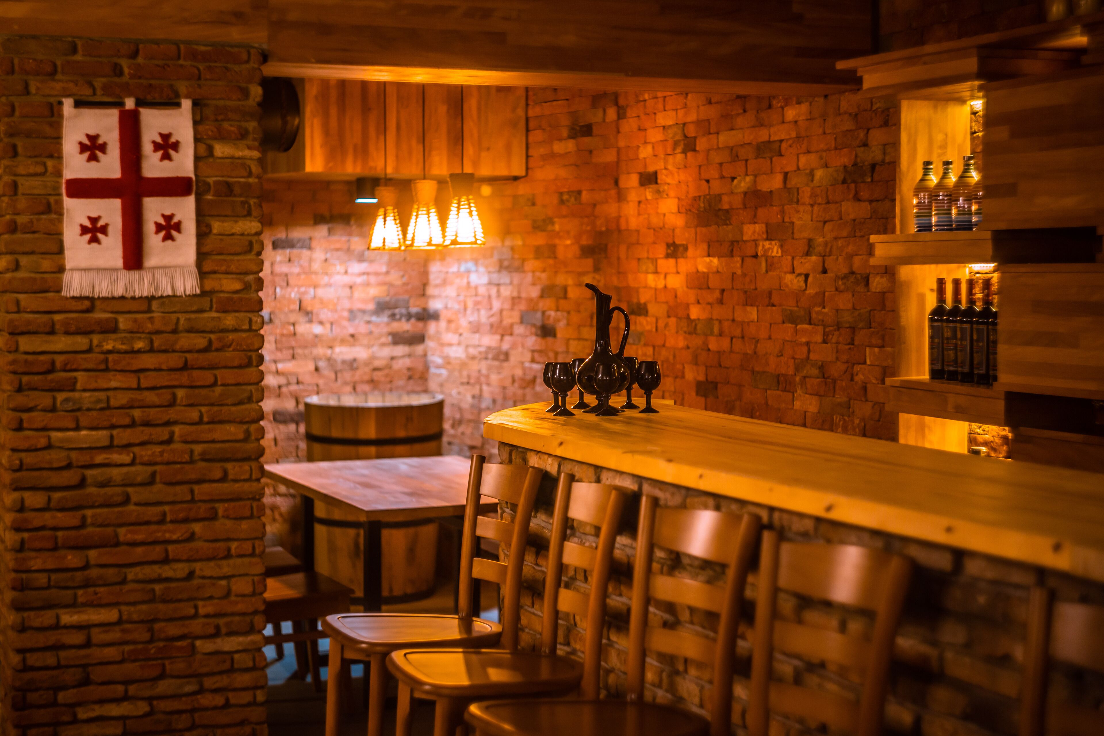 A cozy bar area with a brick wall, wooden bar counter, and bar stools. There are hanging lights above the bar and a cross decoration on the wall.