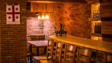 A cozy bar area with a brick wall, wooden bar counter, and bar stools. There are hanging lights above the bar and a cross decoration on the wall.