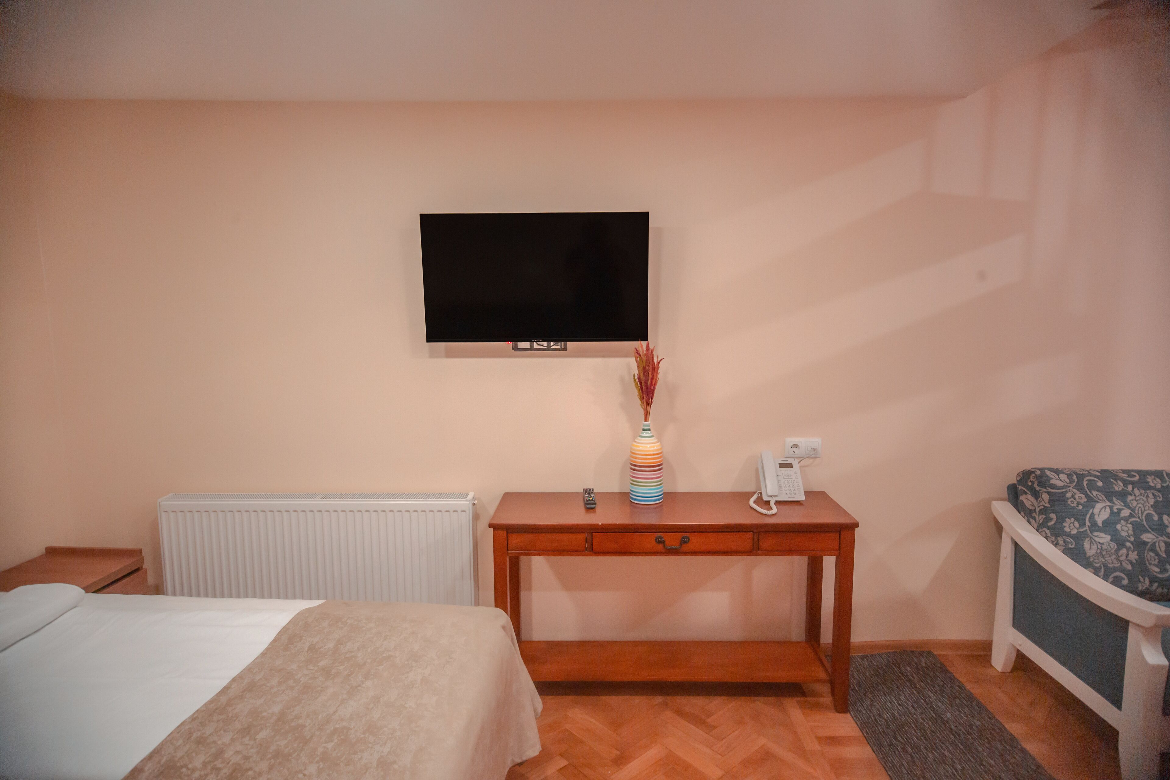 Habitación de hotel con cama, mesita de noche de madera, escritorio con teléfono, radiador y televisor montado en la pared.