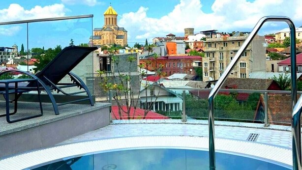 Una piscina en la azotea con barandilla de cristal, una silla de descanso y vistas a un paisaje urbano con un edificio de cúpula amarilla destacada al fondo.