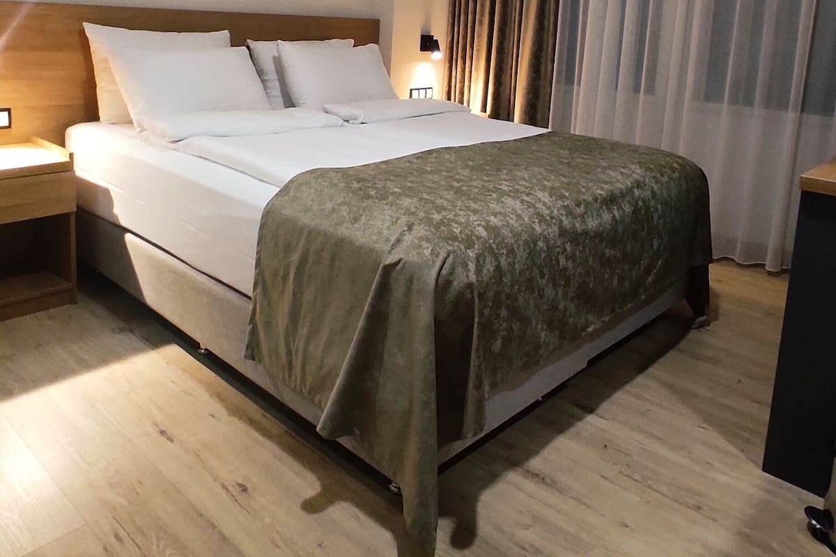 A hotel room with a large bed, wooden flooring, a nightstand, a lamp, and a curtain.