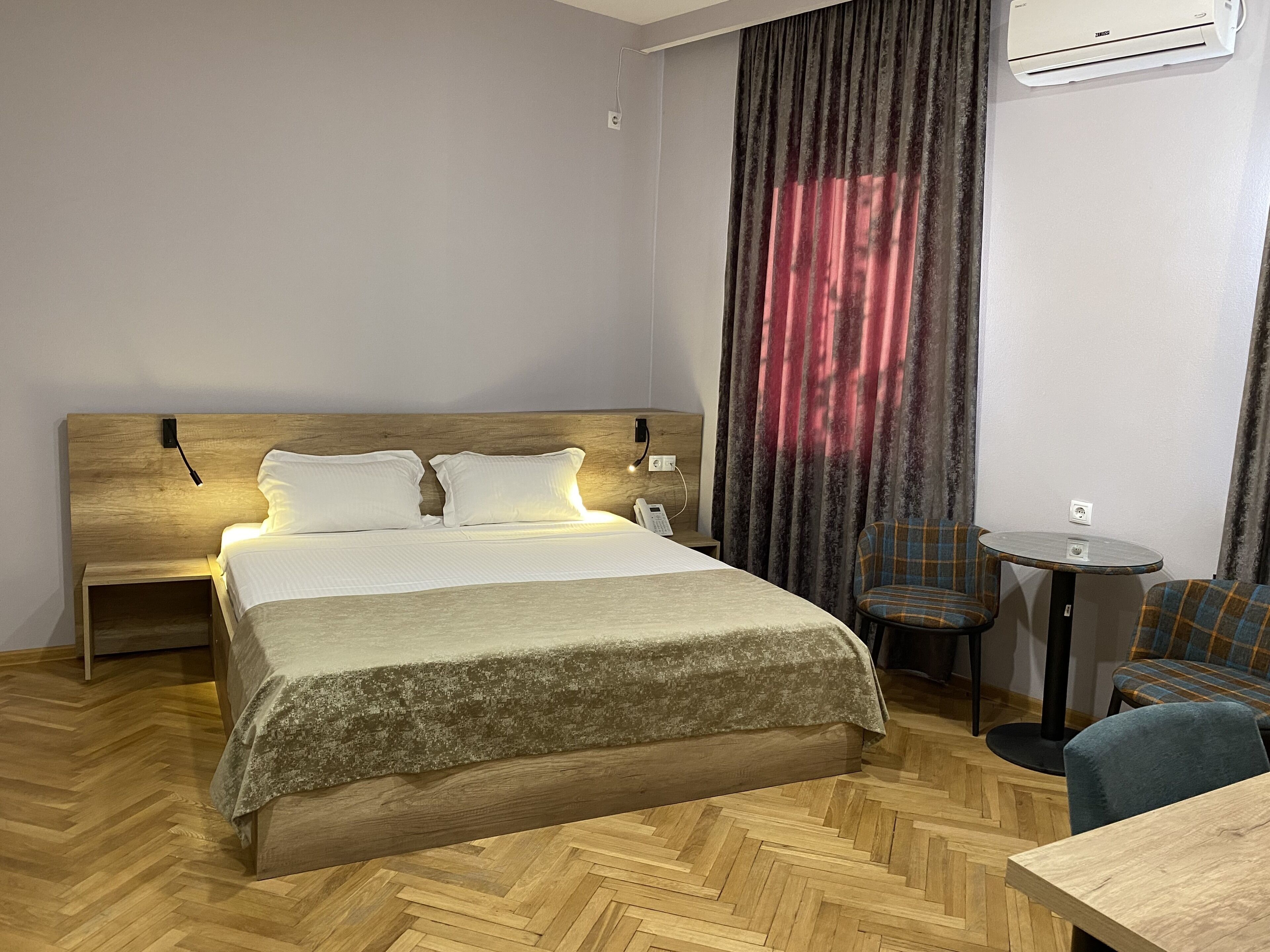 Une chambre d’hôtel avec un lit en bois, une petite table ronde, une chaise et un appareil de climatisation.