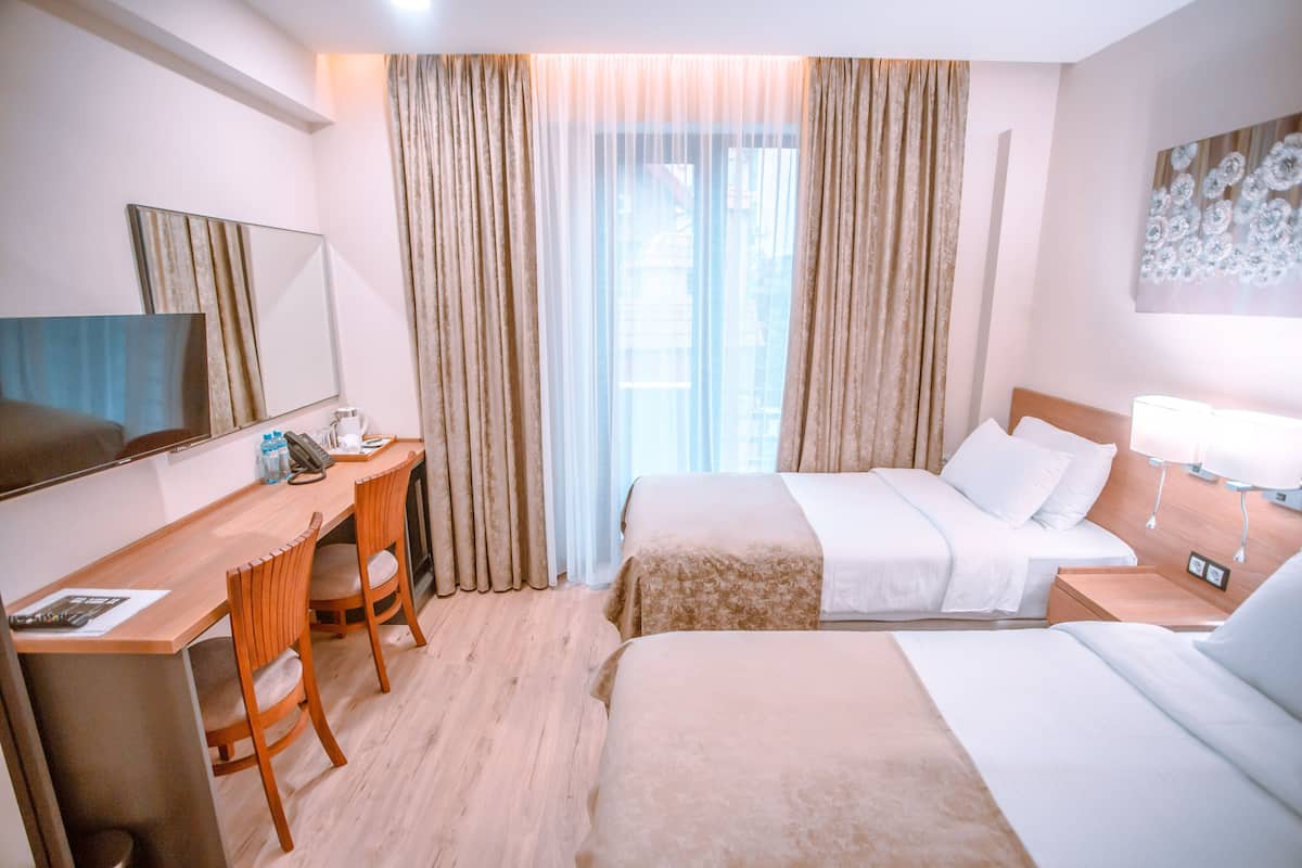 A hotel room with two beds, a desk with a chair, a television, and a large window with curtains.