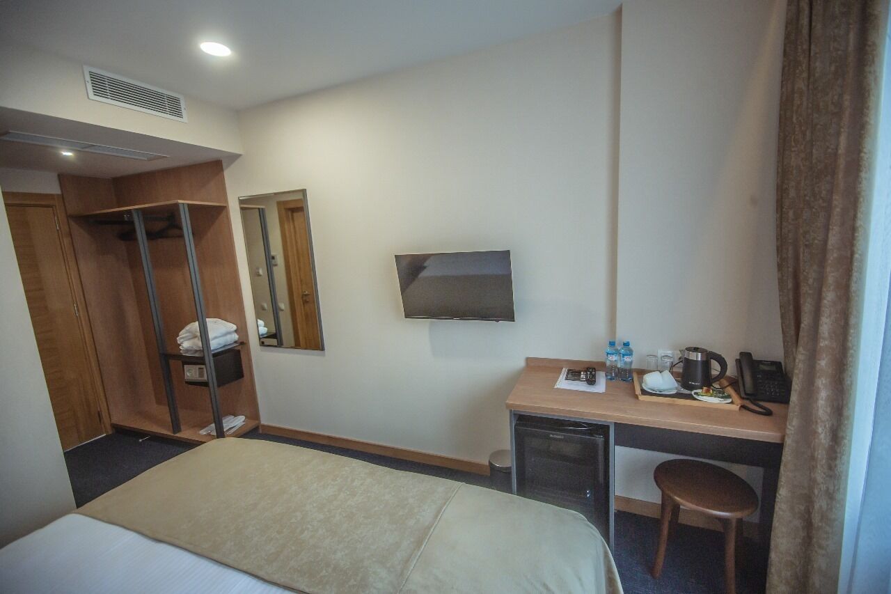 Ein Hotelzimmer mit einem Bett, einem Schreibtisch mit Computer, einem Spiegel, einem Fernseher und einer kleinen Küchenzeile.