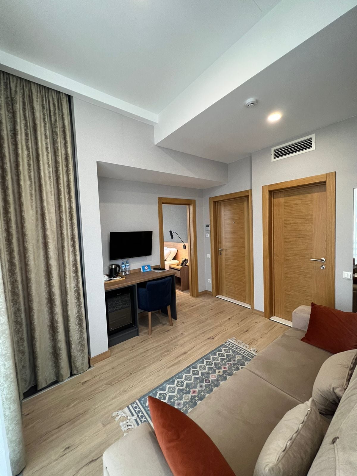 A modern living room with a sofa, a desk, and a television. There are two wooden doors and a window with curtains.