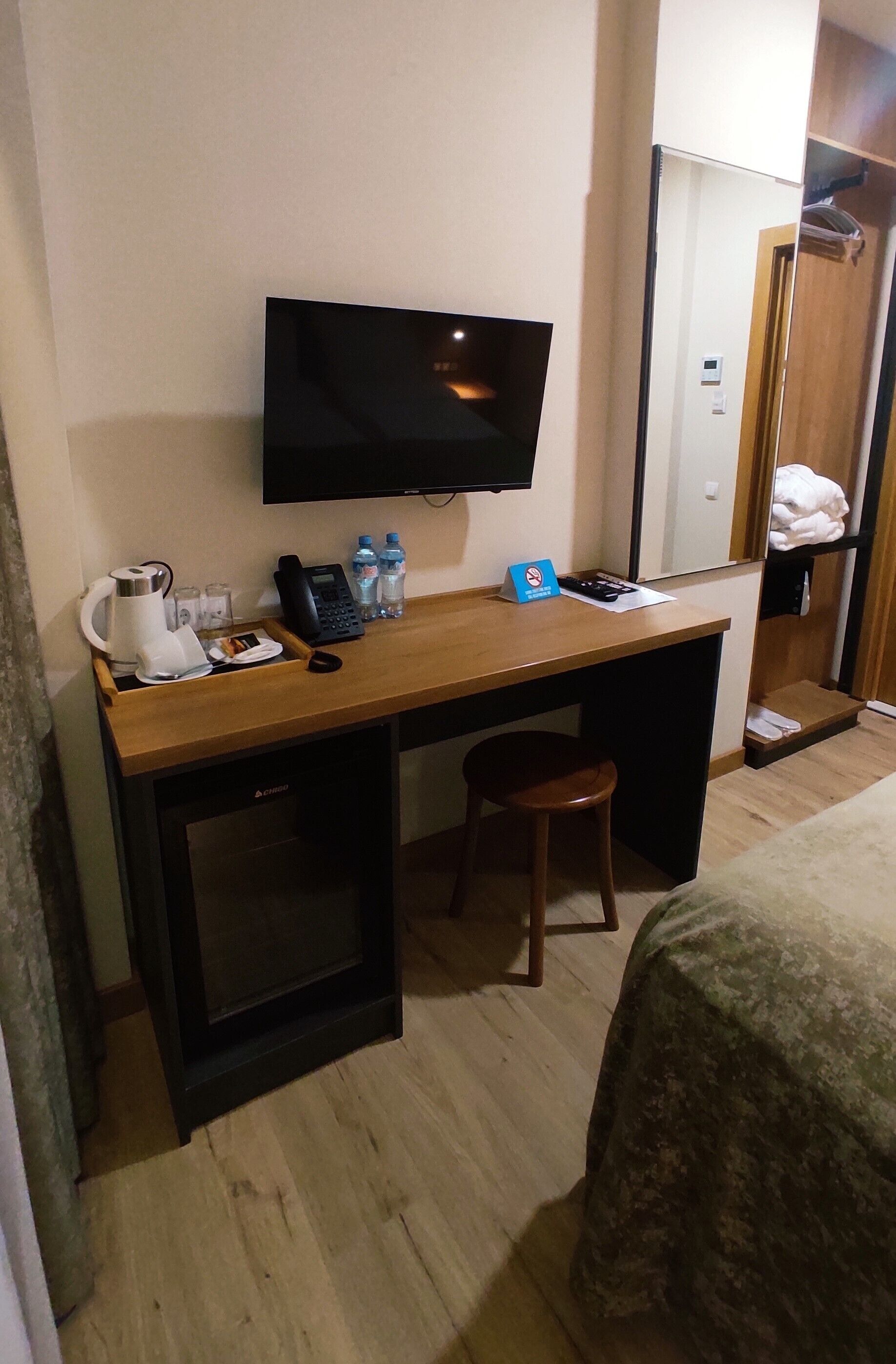 A hotel room with a wooden desk, a television, a telephone, and a small stool.