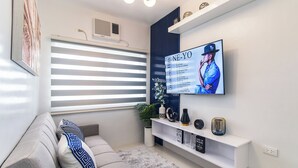 A modern living room with a grey sofa, a mounted TV displaying a music album, a ceiling fan, and a window with blinds.