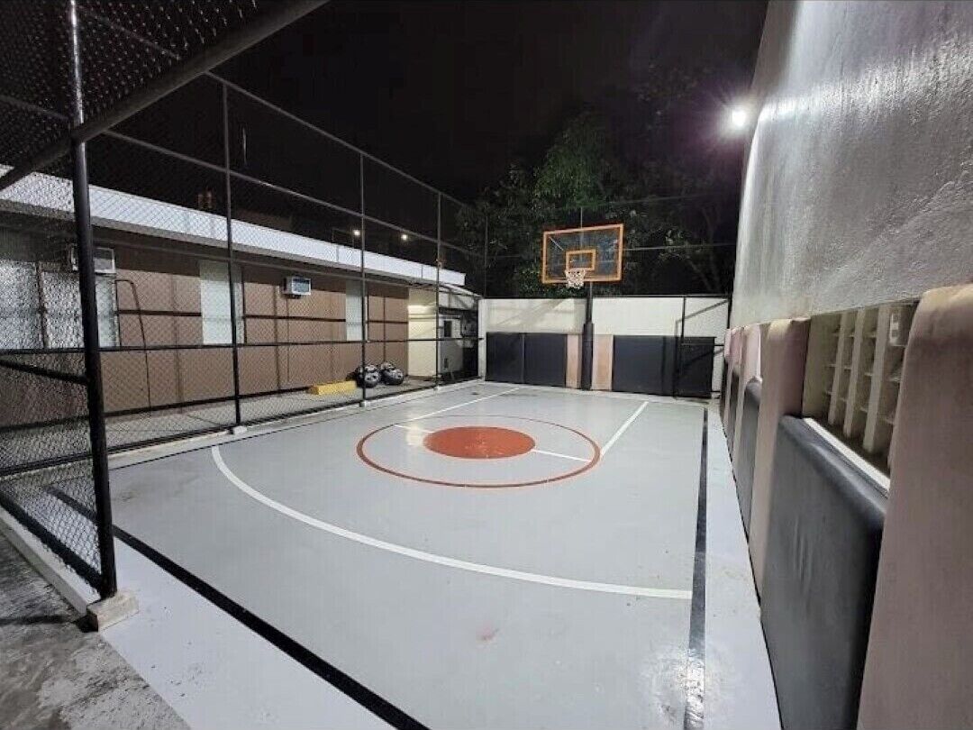 An outdoor basketball court with a metal fence, a basketball hoop, and a bench.