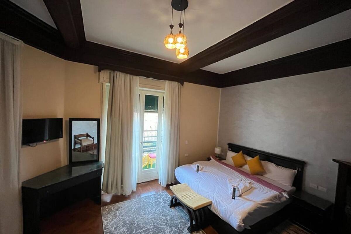 Un dormitorio con cama, mesitas de noche, televisor y ventana con cortinas.
