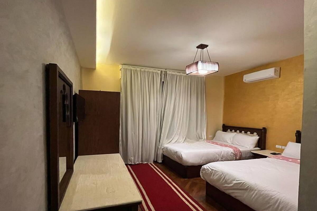 Habitación de hotel con dos camas, un banco, un escritorio, una lámpara colgante y un equipo de aire acondicionado.