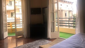 Habitación de hotel con cama, televisión en la pared y balcón con una silla y vistas a edificios.