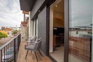 Un balcón con una silla y una mesa, un área de cocina visible a través de una puerta abierta, y vistas de la ciudad.