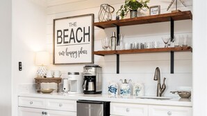 A kitchen with white cabinetry, a coffee maker, a coffee pot, and a sign that reads "The Beach our happy place".