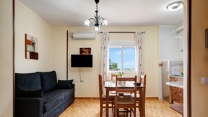 Ein Wohnzimmer mit einer Couch, einem Esstisch mit Stühlen, einem an der Wand montierten Fernseher und einem Fenster mit Vorhängen.