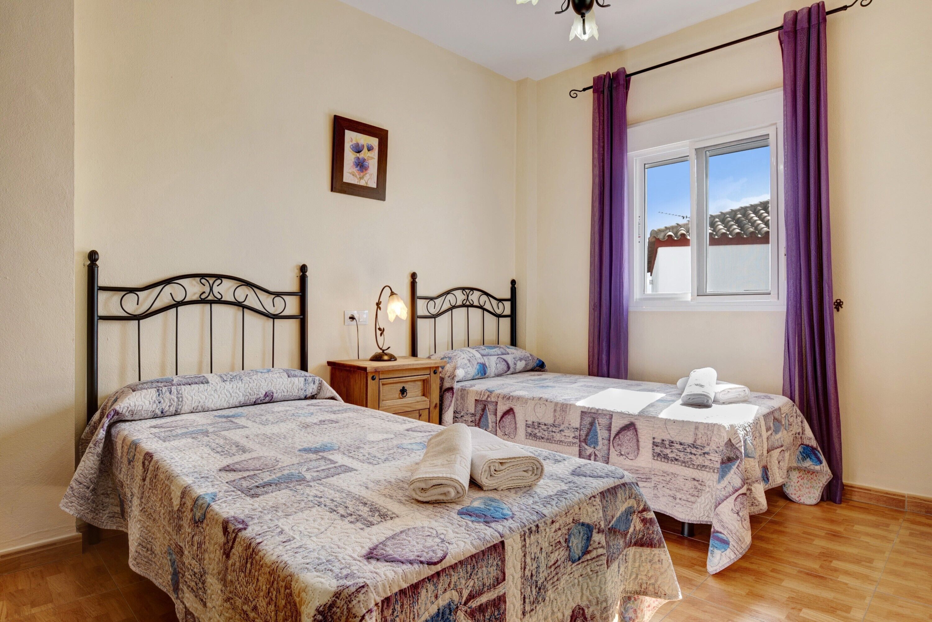 A bedroom with two beds, a nightstand, a table with a towel, a window with purple curtains, and a picture on the wall.