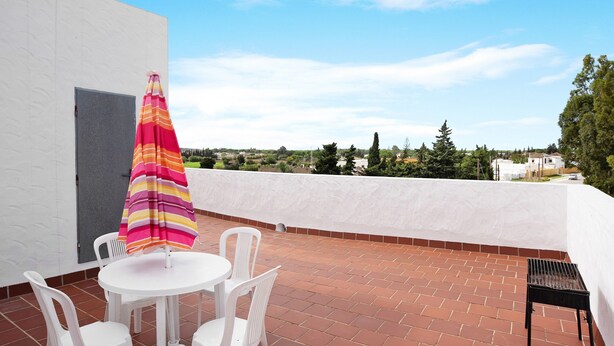 Eine Dachterrasse mit weißem Tisch und Stühlen, einem gestreiften Sonnenschirm und Blick auf die umliegende Landschaft.