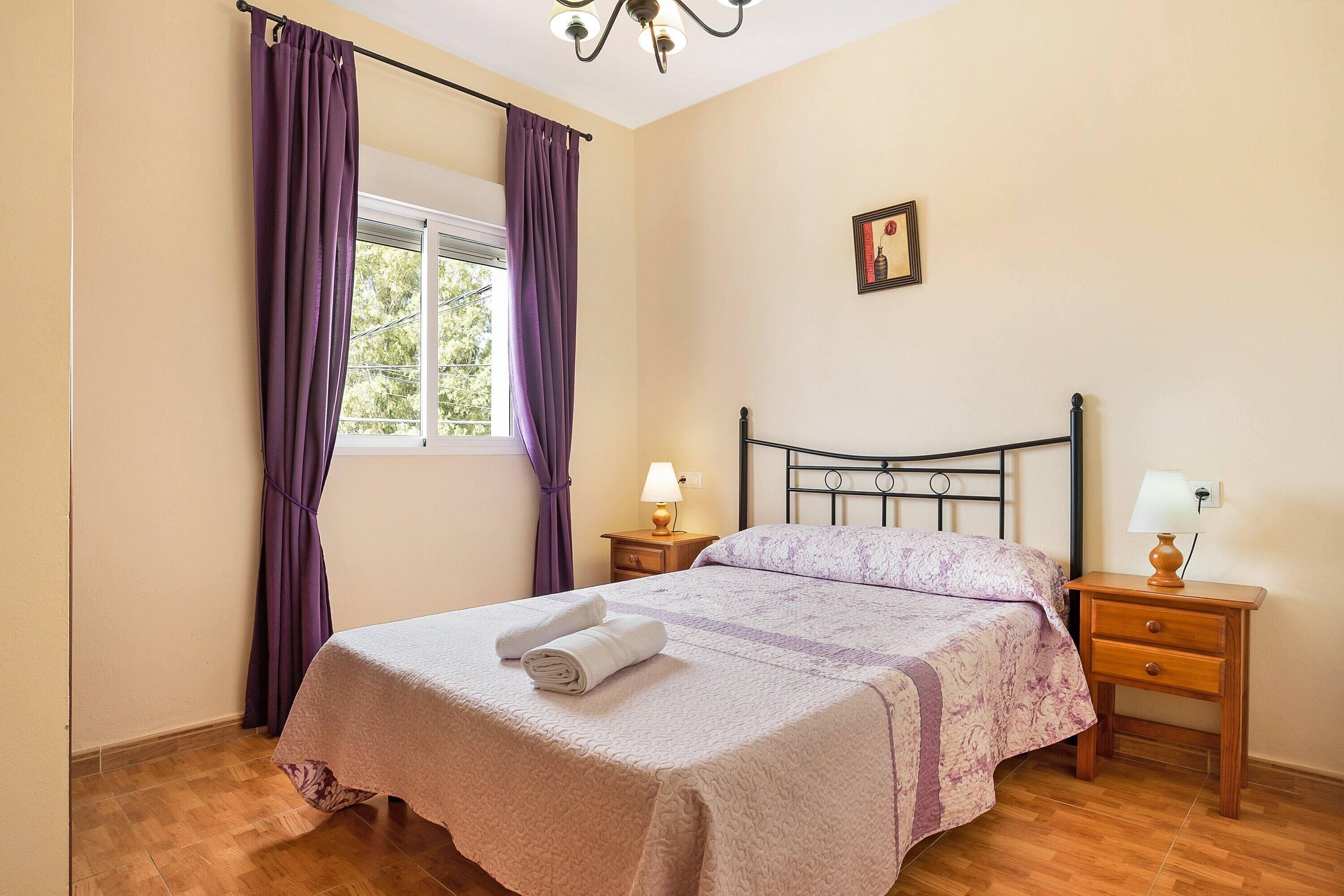 Een slaapkamer met een bed, een raam met gordijnen, een nachtkastje, een lamp en een schilderij aan de muur.