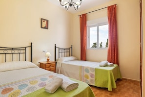 A bedroom with two beds, a window with curtains, a bedside table with a lamp, and a picture on the wall.