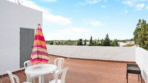 Eine Dachterrasse mit weißer Wand, einem gestreiften Sonnenschirm, einem runden Tisch mit Stühlen und Blick auf Grünflächen und Gebäude.
