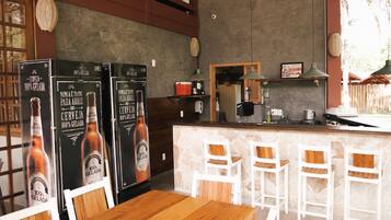 Zona de bar con barra de piedra, taburetes blancos y dos refrigeradores mostrando cerveza '100% Gelada'.
