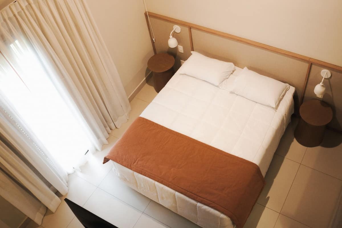 Cama bem arrumada com roupa de cama branca e castanha, duas mesas de cabeceira com abajures e uma janela com cortinas.
