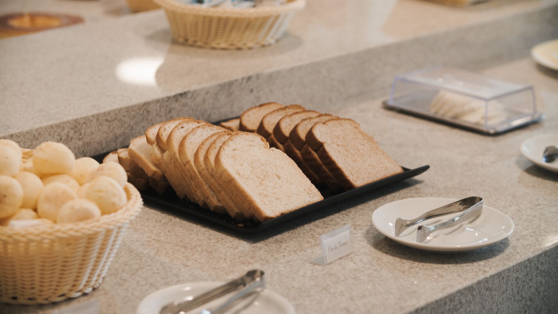 Una barra de buffet con pan en rebanadas, una cesta de pasteles y un recipiente de plástico transparente.